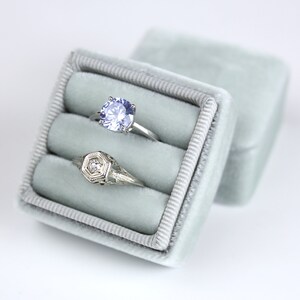 May include: A light gray velvet ring box with two rings inside. One ring has a large, light blue gemstone, and the other is a filigree design with a small center stone. The box is open, with the lid slightly ajar.