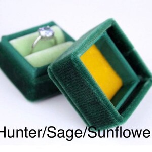 May include: A dark green velvet ring box with a light green interior and a yellow base. A silver ring with a light blue gemstone sits inside the open box. The text "Hunter/Sage/Sunflower" is at the bottom.