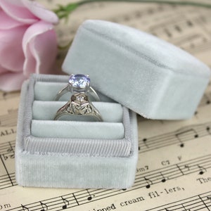 May include: Two rings rest in a light blue velvet ring box. One ring has a silver band with a light blue gemstone, and the other has an ornate silver band. A light blue velvet box lid is open next to the box. A pink rose is in the background.