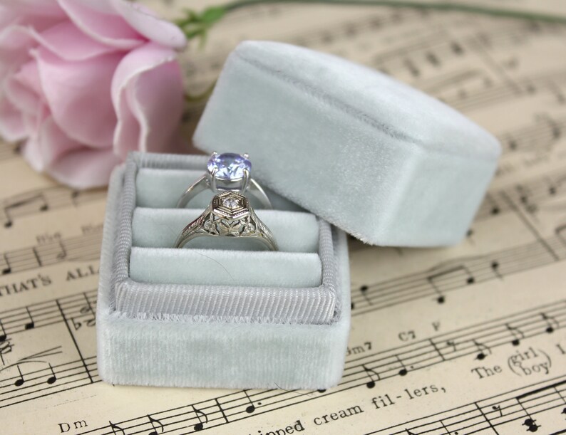 May include: Two rings rest in a light blue velvet ring box. One ring has a silver band with a light blue gemstone, and the other has an ornate silver band. A light blue velvet box lid is open next to the box. A pink rose is in the background.