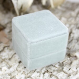 Puede incluir: Una caja de anillo de terciopelo azul claro con una tapa con bisagras. La caja es cuadrada y tiene una textura suave y lisa.