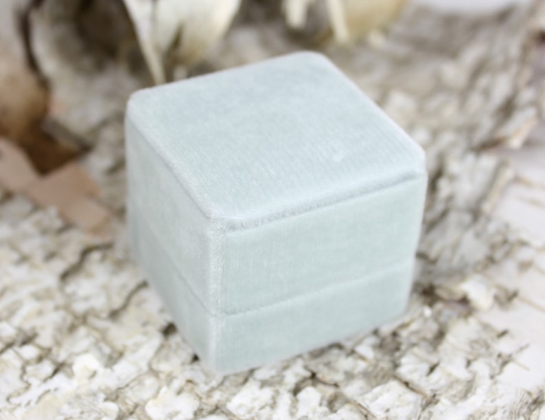 Puede incluir: Una caja de anillo de terciopelo azul claro con una tapa con bisagras. La caja es cuadrada y tiene una textura suave y lisa.