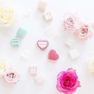 May include: An overhead shot features several ring boxes in various pastel colors, including pink, mint green, and white, scattered among white and pink roses. One box is heart-shaped. The image has a soft, romantic aesthetic.