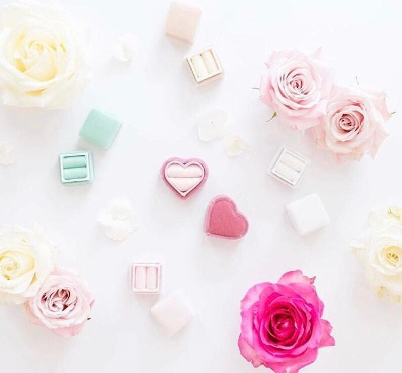 May include: An overhead shot features several ring boxes in various pastel colors, including pink, mint green, and white, scattered among white and pink roses. One box is heart-shaped. The image has a soft, romantic aesthetic.
