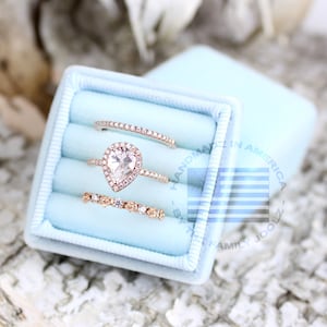 May include: Three rings in a light blue velvet ring box. The center ring features a pear-shaped diamond with a halo setting. The other two rings are thin bands with small diamonds.