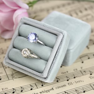 May include: A light blue velvet ring box with two rings inside. One ring has a large, round, light purple gemstone. The other ring has a geometric design with a small, clear gemstone. The box is open, and the lid is next to it.