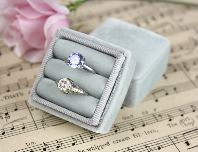 May include: A light blue velvet ring box with two rings inside. One ring has a large, round, light purple gemstone. The other ring has a geometric design with a small, clear gemstone. The box is open, and the lid is next to it.
