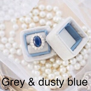 May include: A ring with a large oval blue gemstone and a halo of smaller stones sits in a light blue velvet ring box. The box is inside a white velvet box. The text "Grey & dusty blue" is at the bottom of the image.