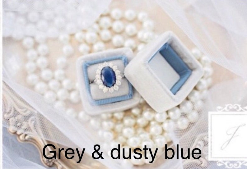 May include: A ring with a large oval blue gemstone and a halo of smaller stones sits in a light blue velvet ring box. The box is inside a white velvet box. The text "Grey & dusty blue" is at the bottom of the image.