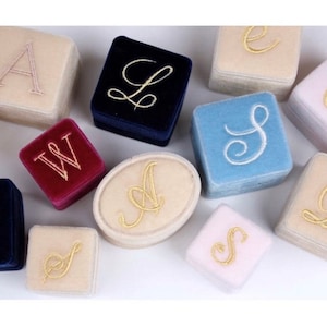 May include: A collection of velvet ring boxes in various colors, including navy blue, light blue, and cream. Each box is embroidered with a different gold letter. The boxes are square and oval-shaped, ideal for jewelry storage or presentation.