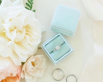 May include: A light blue velvet ring box with a diamond engagement ring inside. The box is surrounded by white and peach roses.
