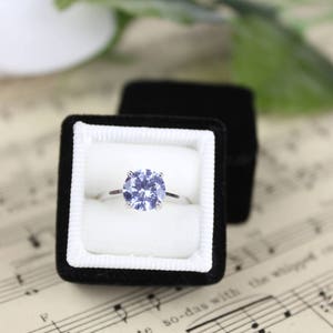 May include: A silver engagement ring with a large, round, light blue gemstone is displayed in an open black velvet ring box. The ring box sits on a sheet of vintage sheet music, with a white flower and green leaves in the background.
