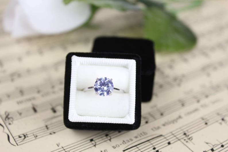 May include: A silver engagement ring with a large, round, light blue gemstone is displayed in an open black velvet ring box. The ring box sits on a sheet of vintage sheet music, with a white flower and green leaves in the background.
