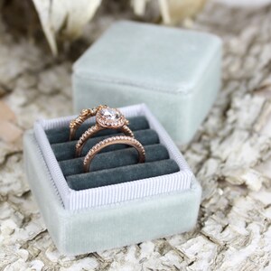 Puede incluir: Una caja de anillo de terciopelo azul claro con tres anillos en su interior. Los anillos son de oro con diamantes. La caja est&aacute; sobre una superficie blanca y marr&oacute;n.