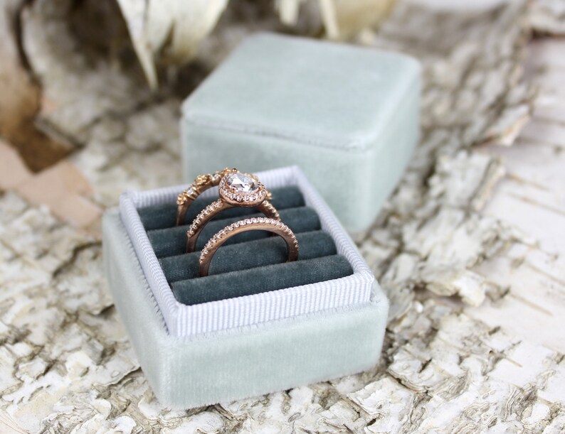 Puede incluir: Una caja de anillo de terciopelo azul claro con tres anillos en su interior. Los anillos son de oro con diamantes. La caja est&aacute; sobre una superficie blanca y marr&oacute;n.