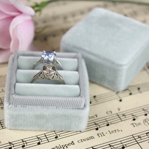 May include: Two rings rest in a light blue velvet ring box, with a matching lid beside it. One ring has a round, light blue gemstone, and the other has an ornate, filigree design. The box sits on sheet music, with a pink rose in the background.