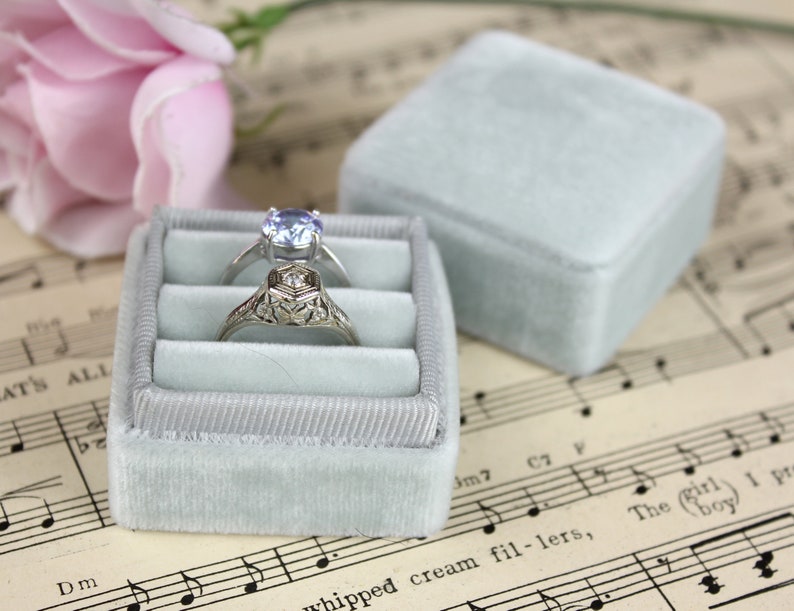 May include: Two rings rest in a light blue velvet ring box, with a matching lid beside it. One ring has a round, light blue gemstone, and the other has an ornate, filigree design. The box sits on sheet music, with a pink rose in the background.