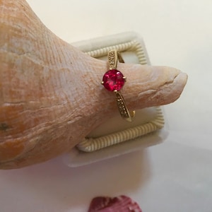 Puede incluir: Un anillo de oro con una piedra preciosa roja y pequeños diamantes en la banda.