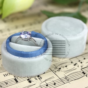 May include: A light blue ring box with a blue ribbon and a silver ring with a purple gemstone. The box is sitting on a sheet of music with the text "HANDMADE IN AMERICA - THE FAMILY JOOLZ."