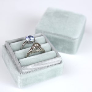 May include: Two rings rest in a light blue velvet ring box. One ring has a round, light blue gemstone, and the other has a hexagonal design. The ring box is open, with the lid slightly behind it.