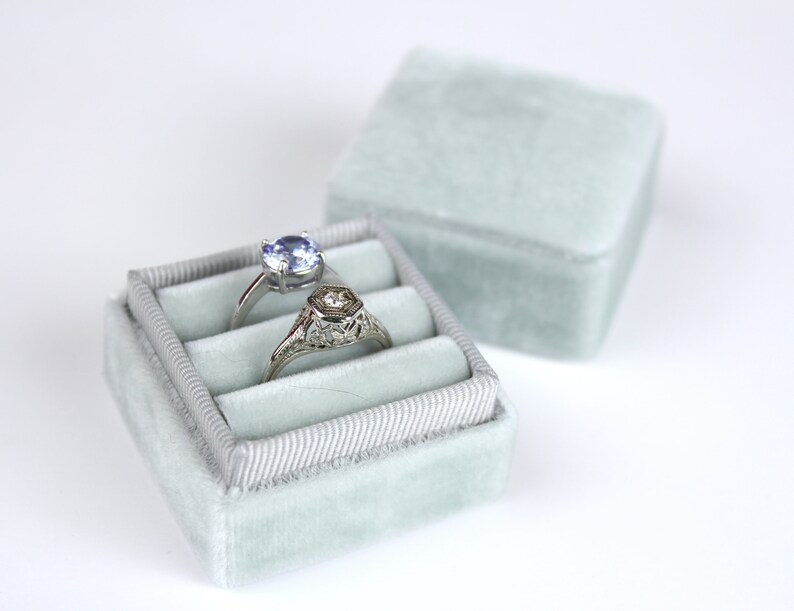 May include: Two rings rest in a light blue velvet ring box. One ring has a round, light blue gemstone, and the other has a hexagonal design. The ring box is open, with the lid slightly behind it.