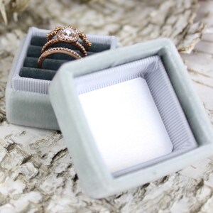 Puede incluir: Una caja de anillo de terciopelo azul claro con un interior blanco. Un anillo de oro con una piedra central grande y piedras m&aacute;s peque&ntilde;as alrededor est&aacute; dentro de la caja.