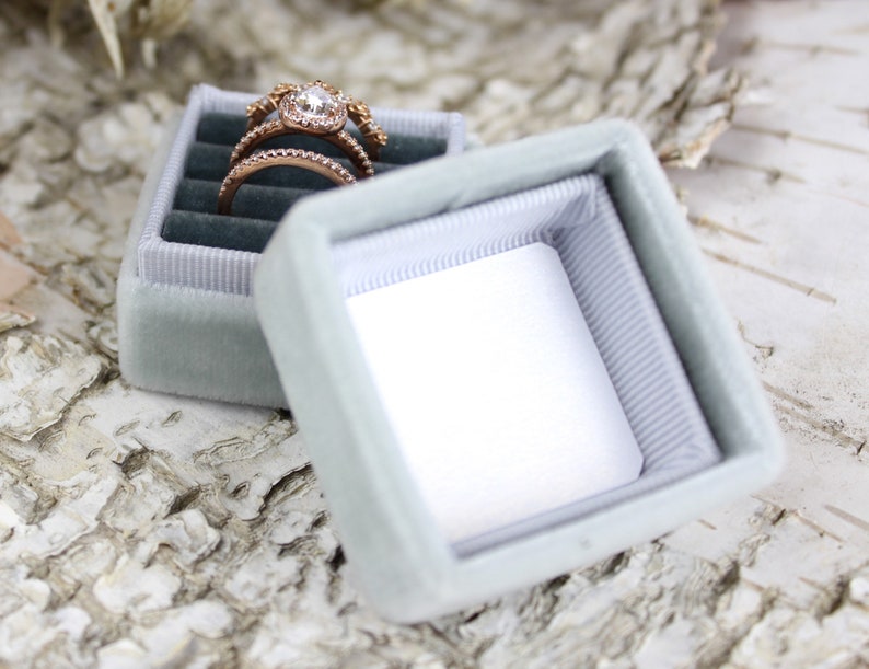 Puede incluir: Una caja de anillo de terciopelo azul claro con un interior blanco. Un anillo de oro con una piedra central grande y piedras m&aacute;s peque&ntilde;as alrededor est&aacute; dentro de la caja.