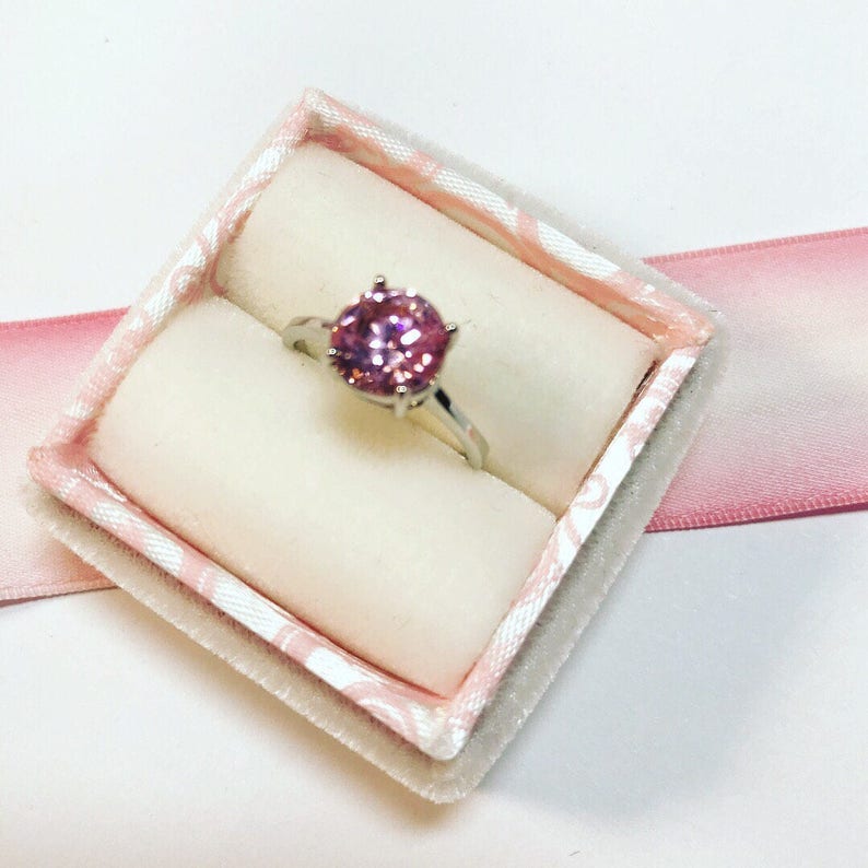 May include: A silver ring with a large, round, pink gemstone is displayed in a pink and white patterned ring box. The ring box is lined with a soft, white material and sits on a white surface with a pink ribbon.