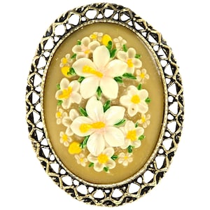 May include: An oval brooch with a floral design. The brooch has a cream-coloured background with white and yellow flowers and green leaves. It is framed by a gold-coloured filigree border, adding a vintage touch to the piece.
