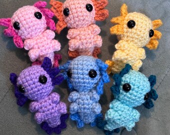 May include: Six crocheted axolotl plushies in different colors: pink, orange, yellow, light blue, purple, and blue. The axolotls are all facing forward with their eyes closed.