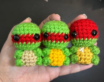 Könnte beinhalten: Drei handgefertigte Schildkrötenfiguren aus Häkelarbeit. Jede Schildkröte hat einen grünen Kopf, eine rote Maske und schwarze Augen. Die Schildkrötenschilde sind beige, gelb und grün. Die Figuren werden in einer Hand gehalten.