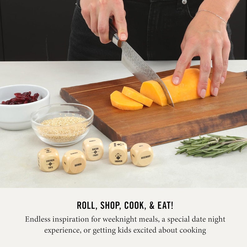 May include: Wooden dice with different food categories printed on each side, such as chicken, tofu, bacon, and quinoa. The dice are on a wooden cutting board with a knife, a bowl of cranberries, a bowl of quinoa, and a sprig of rosemary.