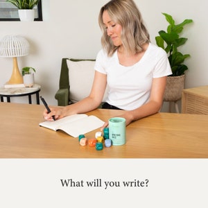 May include: A person is writing in a notebook with a set of colorful writing dice. The dice are in a green container with the words "Writing Dice" on it.