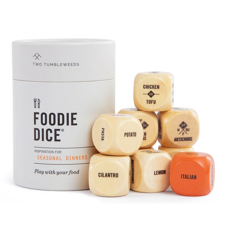 May include: A set of wooden dice with various food-related words printed on each side, including "Pasta", "Potato", "Chicken or Tofu", "Artichoke", "Cilantro", "Mushrooms", "Lemon", and "Italian". The dice are arranged in a pyramid shape on a white surface. The dice are part of a game called "Foodie Dice" and are meant to inspire seasonal dinners.