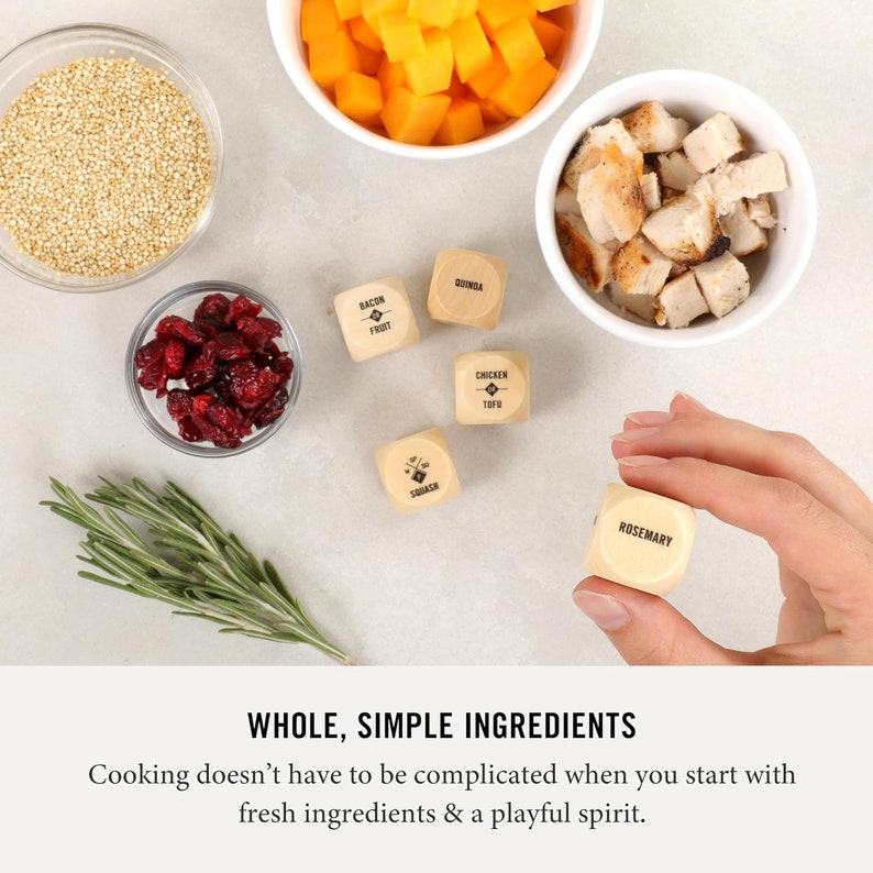 May include: Wooden dice with different food ingredients printed on each side, such as bacon, fruit, squash, chicken tofu, quinoa, and rosemary. The dice are on a white surface with bowls of food ingredients, including cranberries, quinoa, chicken, and butternut squash.