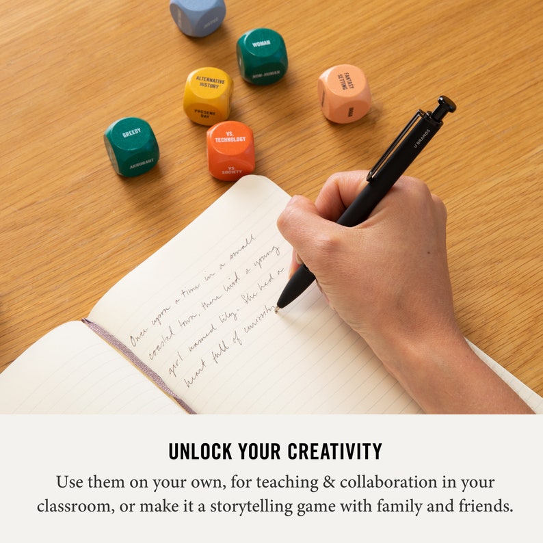 May include: A hand writes in a notebook with a black pen.  The notebook is open to a blank page with lined paper.  There are colorful dice with words on them scattered on a wooden table.  The dice have words like "Greedy", "Technology", "Alternative History", "Setting", "Woman", and "Fantasy".