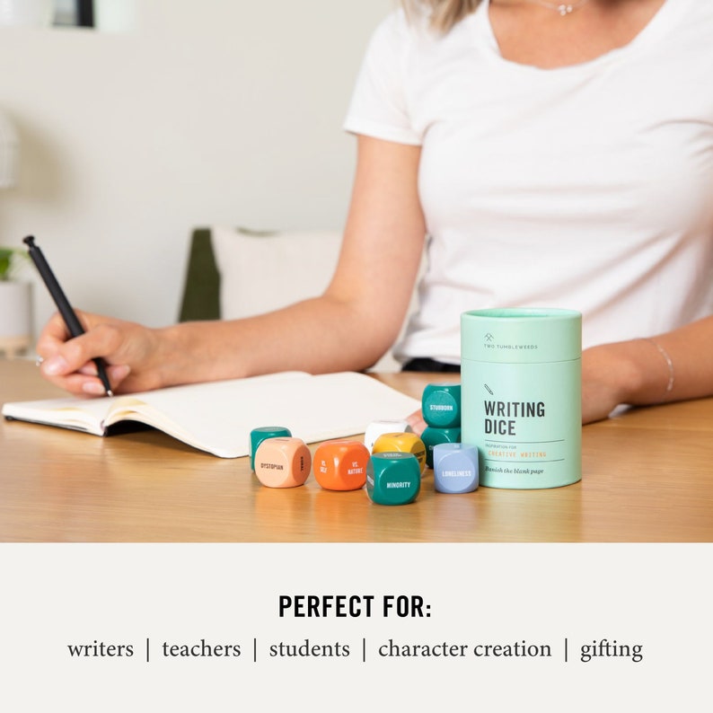 May include: A set of colorful dice with words printed on each side, contained in a green cylinder with the words "Writing Dice" printed on it. The dice are on a wooden table next to a notebook and a pen.