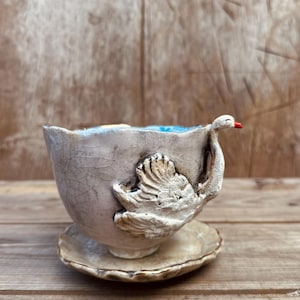 May include: A handcrafted ceramic teacup and saucer set with a crackled off-white glaze. The cup features a three-dimensional sculpted white swan attached to the side, with a small red beak detail. The interior of the cup is painted light blue.