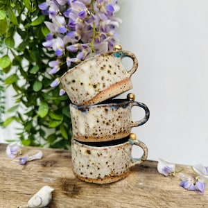 May include: Three stacked ceramic mugs with a speckled beige and brown glaze, each with a handle and a gold accent. The mugs are on a wooden surface, with purple flowers and greenery in the background. The mugs are handmade.