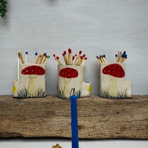 May include: Three ceramic match holders with red mushroom designs and colourful matches. A blue candle stands in a bird-shaped candle holder with a gold crown. The items are on a wooden surface.