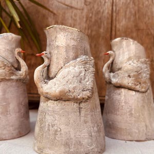 May include: Three conical ceramic pitchers with a weathered, light beige finish. Each pitcher features a sculpted swan relief on the front with a distinct orange beak. The rims of the pitchers have a metallic gold-toned edge, and the bodies have a textured, matte appearance.