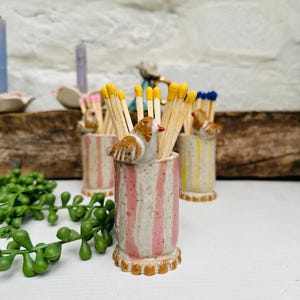 May include: A ceramic matchstick holder with a pink and white striped design. A small bird figurine is perched on the top of the holder. The holder is filled with wooden matchsticks with yellow tips.