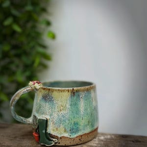 May include: A ceramic mug with a speckled glaze in shades of blue, green, and cream. The mug features a small, sculpted green boot and a red accent near the base, with a tiny white and red mushroom detail attached to the top of the handle.