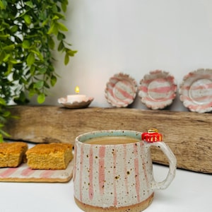 Puede incluir: Una taza de cerámica moteada con rayas verticales rosas, llena de una bebida marrón claro. Un pequeño adorno rojo y dorado está sujeto al asa. Tres platos pequeños a juego con rayas rosas están en el fondo.