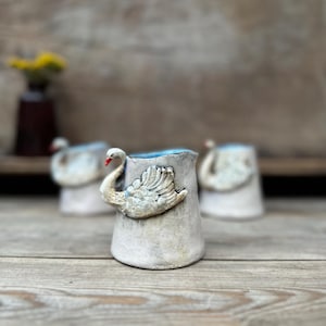 May include: A set of three ceramic pitchers with a textured off-white finish and a blue interior. Each pitcher features a sculpted swan relief on the front with a distinct orange beak. The pitchers have a tapered cylindrical shape and a handle.