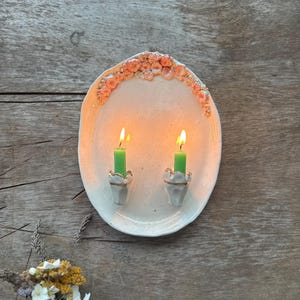 May include: An oval ceramic tray with a speckled off-white finish, featuring a decorative arch of small pink and orange sculpted flowers along the top edge. Two white candle holders with gold rims are attached, each holding a lit green taper candle.