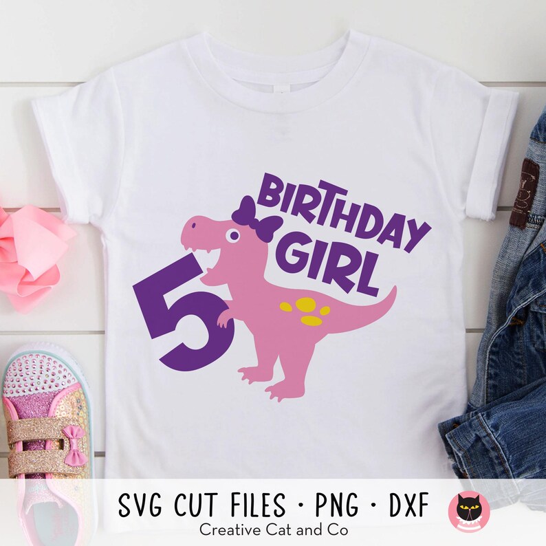 K&ouml;nnte beinhalten: Wei&szlig;es T-Shirt mit einer lila Zahl 5, einem rosa Dinosaurier mit einer lila Schleife und dem Text "Birthday Girl" in lila.