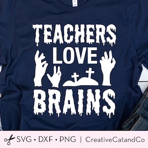 K&ouml;nnte beinhalten: Ein dunkelblaues T-Shirt mit einem wei&szlig;en Grafikdesign, auf dem "Teachers Love Brains" mit Zombie-H&auml;nden und einem Friedhof steht.