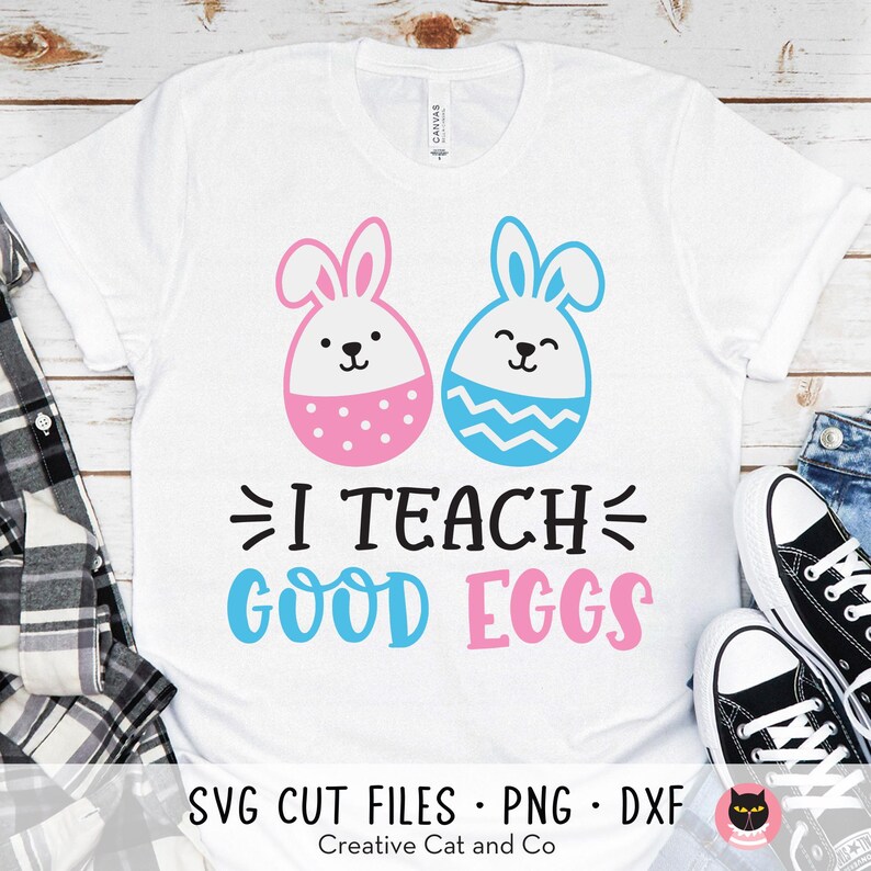 May include: White t-shirt featuring a blue and pink bunny design. The bunnies are holding eggs. The text "I Teach Good Eggs" is printed on the shirt.