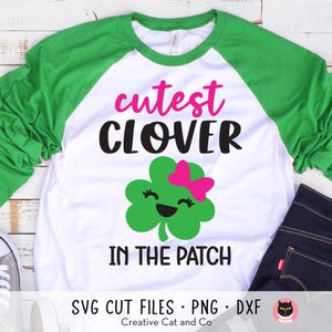 May include: A white and green raglan t-shirt with the text "cutest clover in the patch" in black and pink lettering. A green four-leaf clover with a pink bow and a smiling face is in the centre of the shirt.